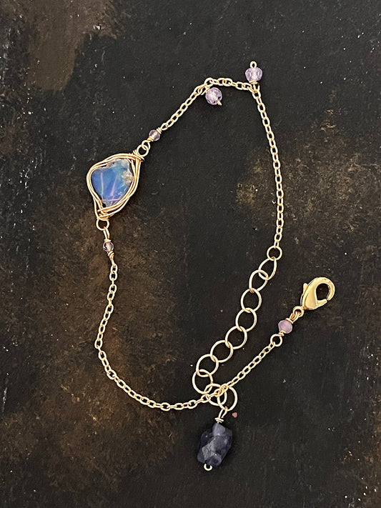 Top-down view of a raw blue opal wire-wrapped bracelet on a dark, textured slate surface, showing the intricate gold chain and purple accent stones.