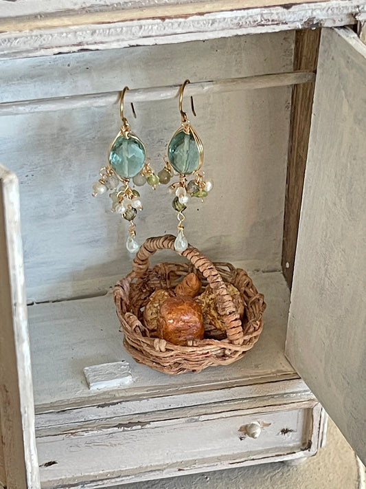 Close up of green quartz and gemstone chandelier earrings hanging inside a miniature rustic white wooden cabinet above a tiny wicker basket. Artisan jewelry handmade by a Japanese designer in Geneva.