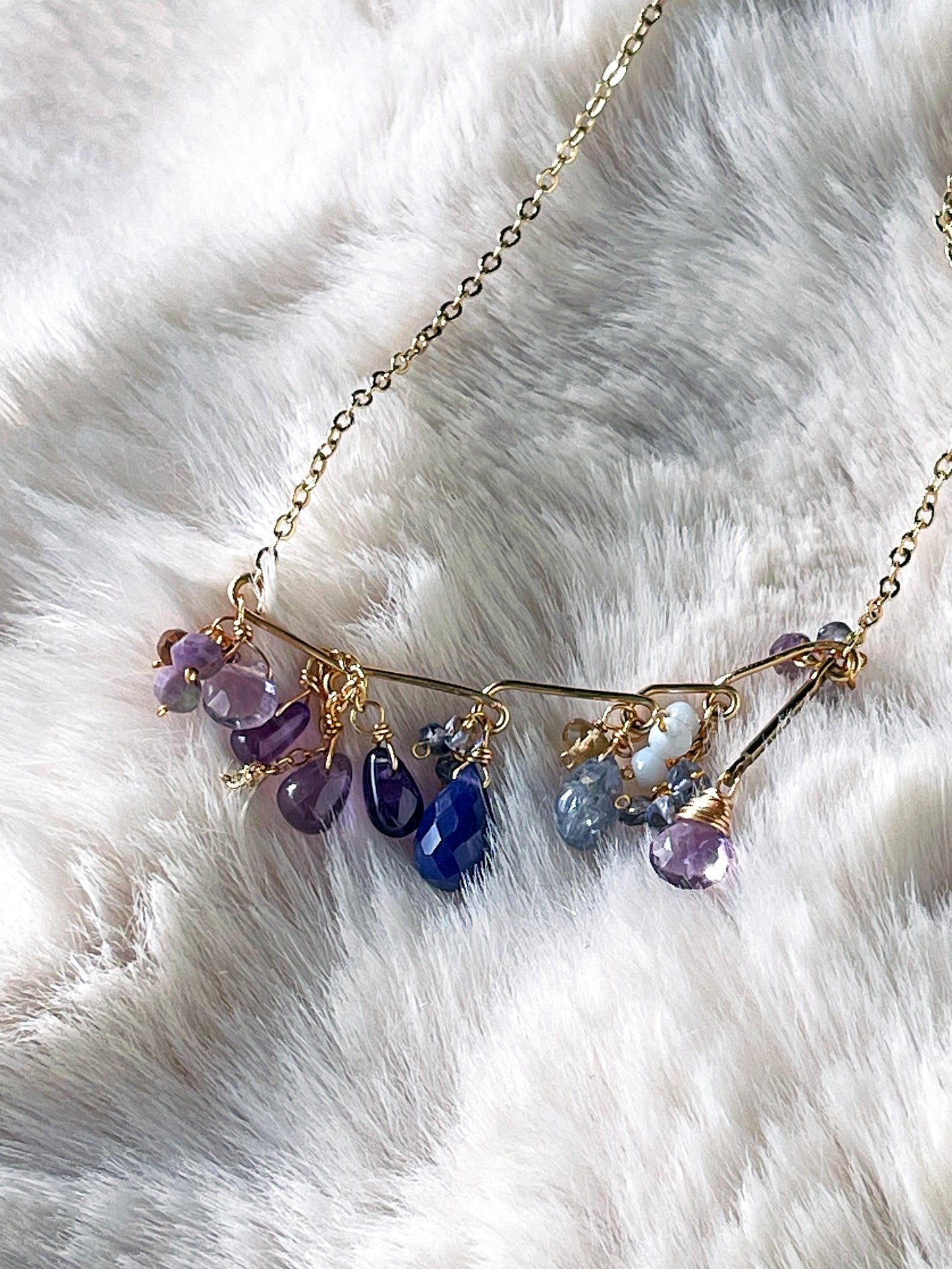 Close-up of unique gold bar necklace with dangling purple chalcedony, amethyst, and tanzanite teardrops resting on a soft white faux fur surface.