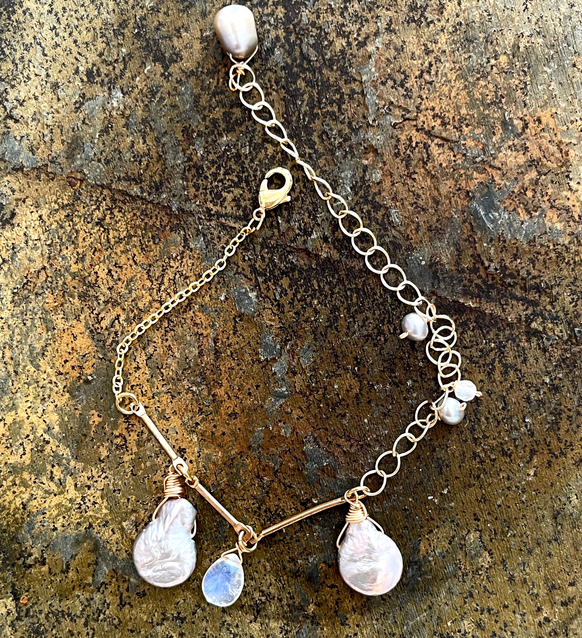 A gold bracelet by LuLu Jewel Design featuring large gray Keshi pearls and a rainbow moonstone charm, displayed on a natural stone surface.