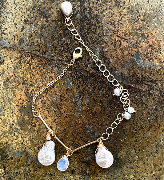 A gold bracelet by LuLu Jewel Design featuring large gray Keshi pearls and a rainbow moonstone charm, displayed on a natural stone surface.