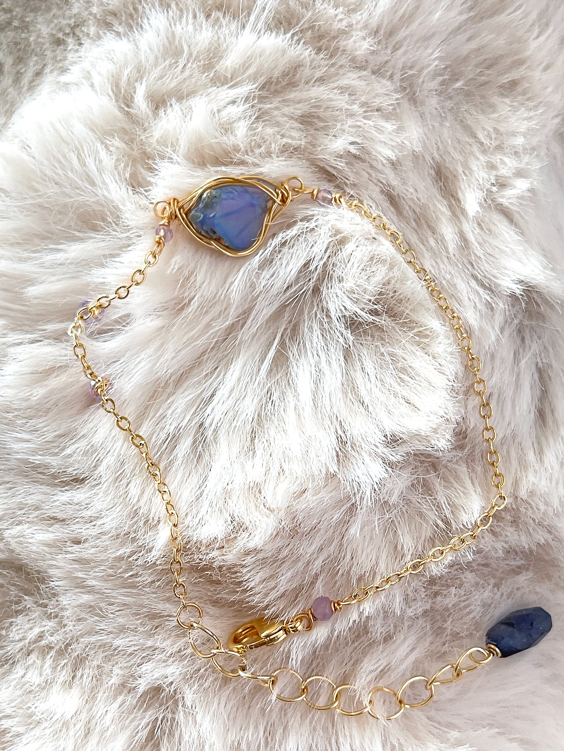 A gold bracelet by LuLu Jewel Design featuring a raw, wire-wrapped blue opal stone on a soft white faux fur background.