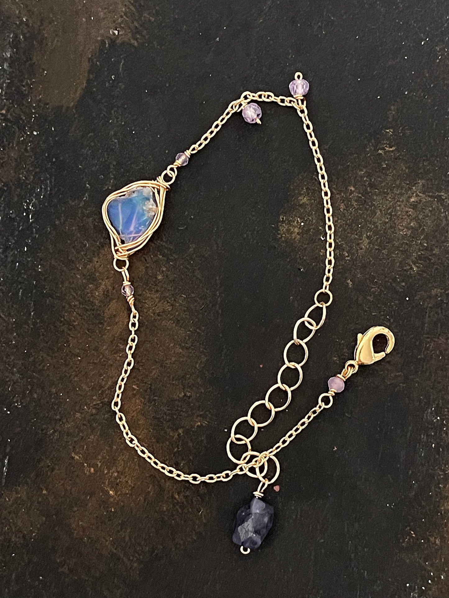 Top-down view of a raw blue opal wire-wrapped bracelet on a dark, textured slate surface, showing the intricate gold chain and purple accent stones.