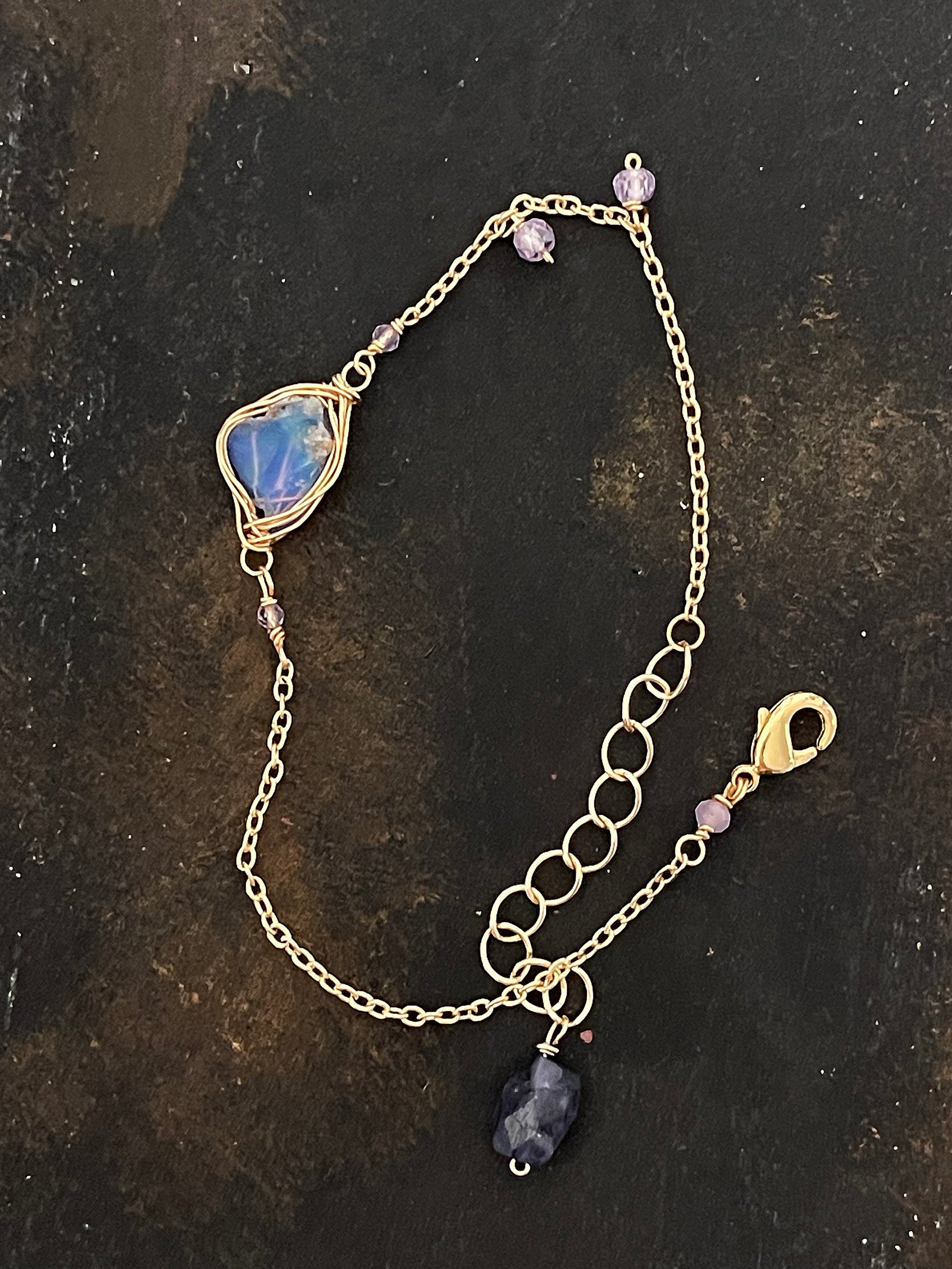 Top-down view of a raw blue opal wire-wrapped bracelet on a dark, textured slate surface, showing the intricate gold chain and purple accent stones.
