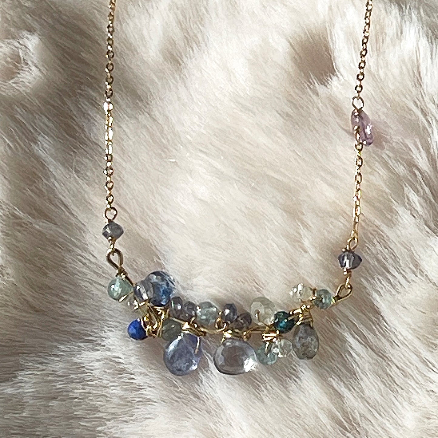 Close-up of a gold necklace from LuLu Jewel Design featuring a cluster of blue gemstones like tanzanite and kyanite on a soft white fur background.