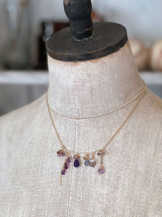 A bar-style necklace by LuLu ewelDesign featuring a row of purple gemstones including amethyst and tanzanite, displayed on a vintage-style mannequin torso.