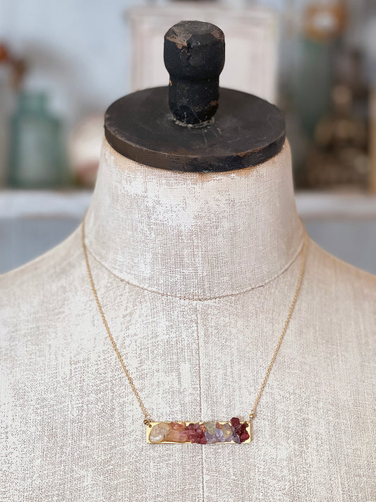 Gold necklace with a bar plate pendant featuring raw garnet, tanzanite, pink tourmaline and moonstone on a antique style mannequin bust.