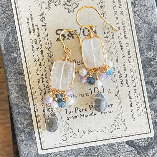 A pair of intricate gold wire-wrapped earrings featuring clear rectangular rutile quartz and grey labradorite drops, displayed on a vintage-style French soap label with "SAVON" text.
